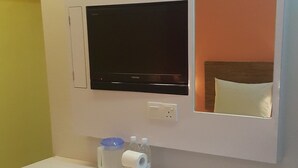 Una habitación de hotel pequeña con un televisor montado en la pared, una mesita blanca con artículos de tocador y un cubo de basura.