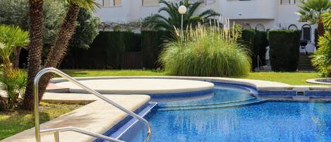 A swimming pool with a metal ladder, surrounded by palm trees and a building with arched windows.