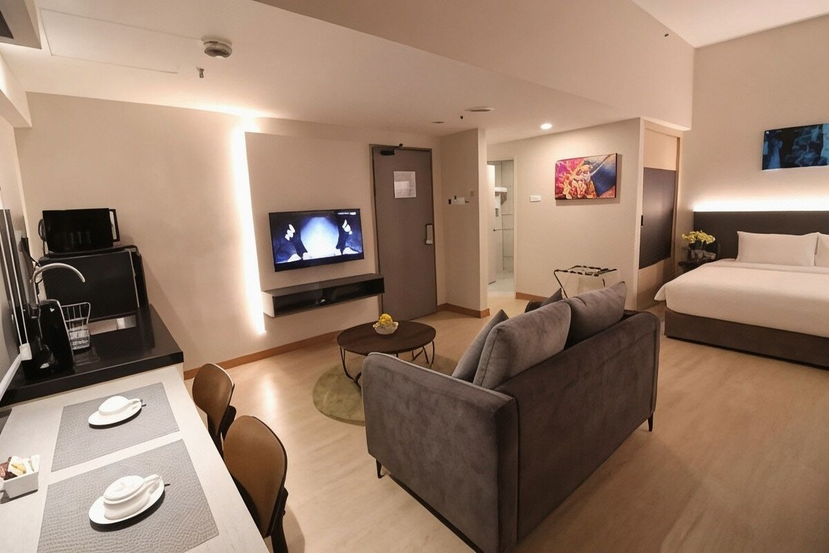 A modern hotel room with a flat-screen TV, a sofa, a bed, and a dining area with a table and chairs.