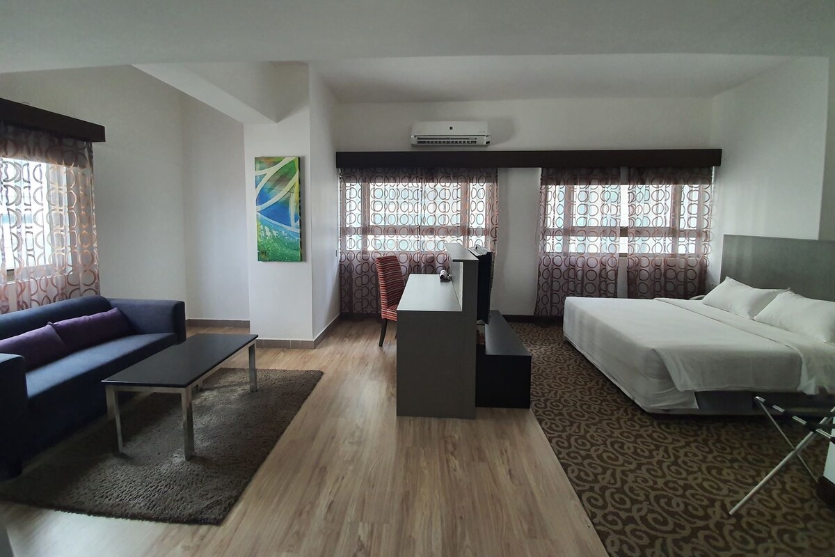 A hotel room with a bed, a sofa, a desk, and a chair. There is a window with curtains, a painting on the wall, and an air conditioning unit.