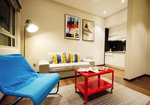 Un salon moderne avec un fauteuil bleu, une table basse rouge, un canapé agrémenté de coussins colorés et deux tableaux abstraits accrochés au mur.