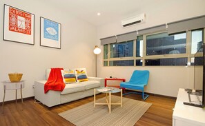A modern living room with a white sofa, colorful cushions, a blue armchair, a small round table, and a red side table. There are two framed artworks on the wall, a window with a view of buildings, and an air conditioning unit above the window. - Crown Regency Serviced Suites (Kuala Lumpur)