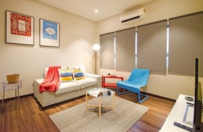 A living room with a sofa, a blue chair, a small table, and a TV. There are two framed artworks on the wall, a basket on a stool, and a window with blinds. - Crown Regency Serviced Suites (Kuala Lumpur)