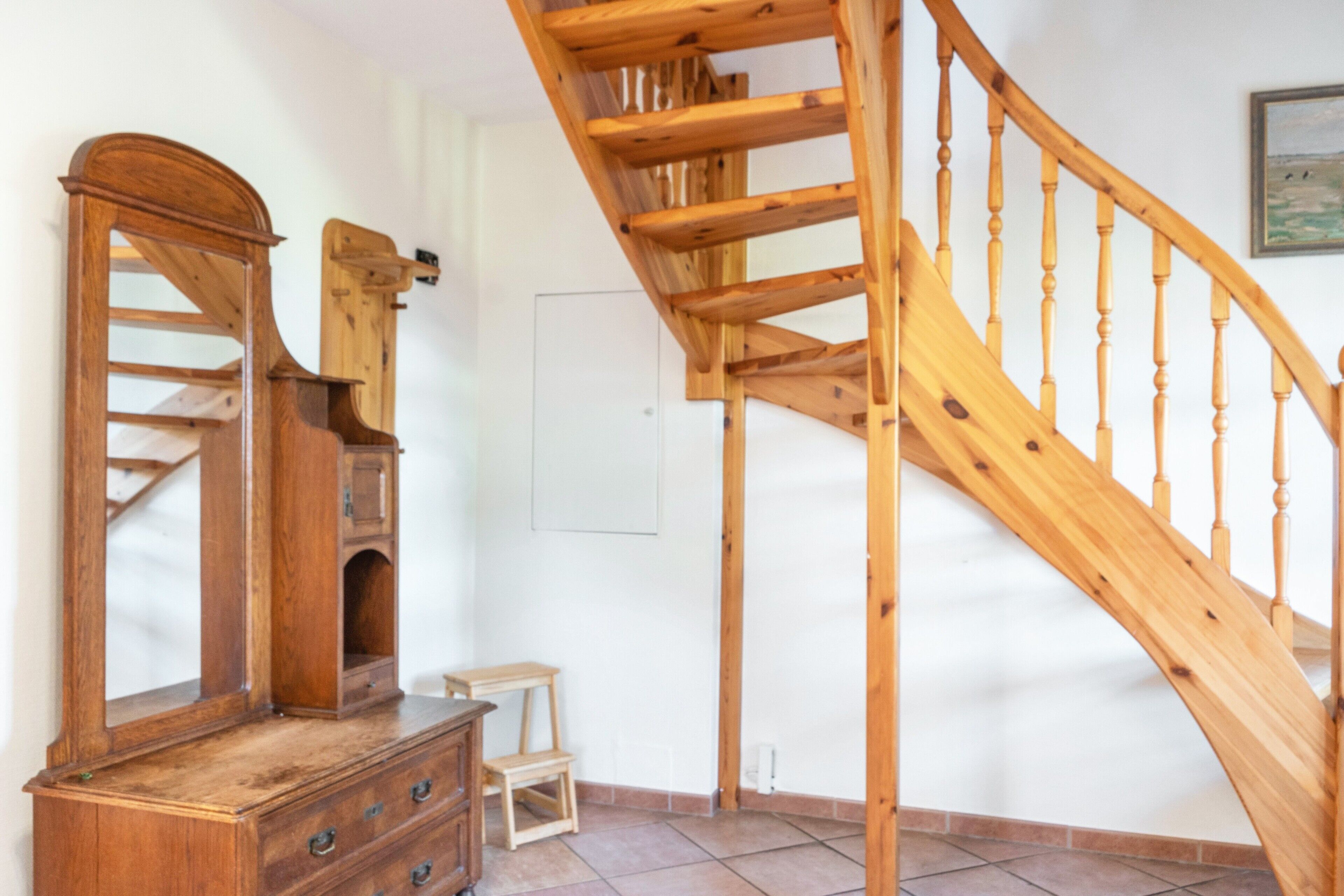 Una escalera de madera con barandilla, un aparador de madera y una mesita en una habitación con paredes blancas y suelo de baldosas.
