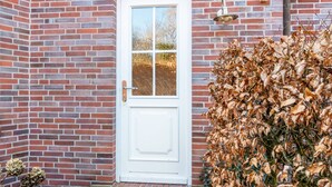 Eine weiße Tür mit Glasfenster, eine Ziegelwand und ein Busch mit braunen Blättern.