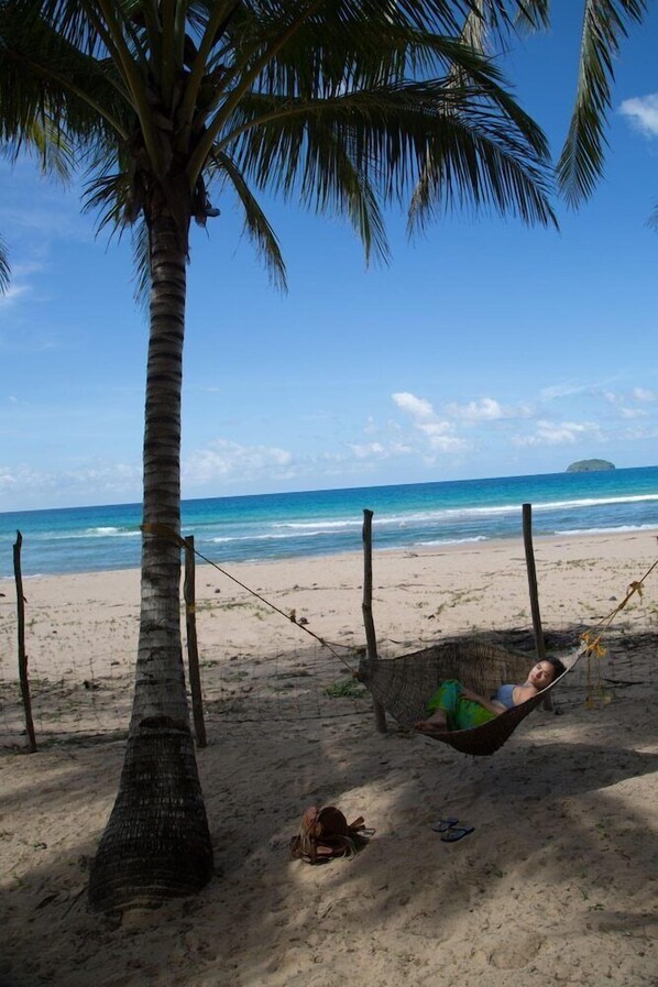 Eine Strandansicht mit einer Hängematte, die an einer Palme befestigt ist, einer Person, die darin entspannt, und einem sandigen Strand mit dem Ozean im Hintergrund.