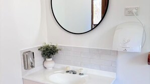 Un baño con un lavamanos blanco, un espejo redondo, una planta en maceta y un purificador de agua.