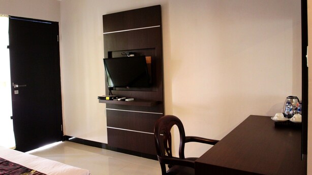 A hotel room with a flat-screen TV mounted on a dark wooden wall, a dining table with a chair, and a bed with a patterned bedspread. - Mansu Hotel & Spa (Denpasar)