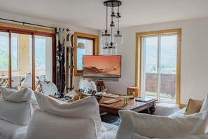 Een woonkamer met een televisie, ramen met houten kozijnen en uitzicht op de omgeving.