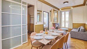 Un comedor con una mesa grande dispuesta para seis personas, rodeada de sillas. Hay un sofá y una mesita con un jarrón de flores.