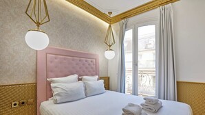 Una cama bien hecha con sábanas y almohadas blancas, un cabecero tapizado en rosa, y vistas a un balcón a través de un gran ventanal.