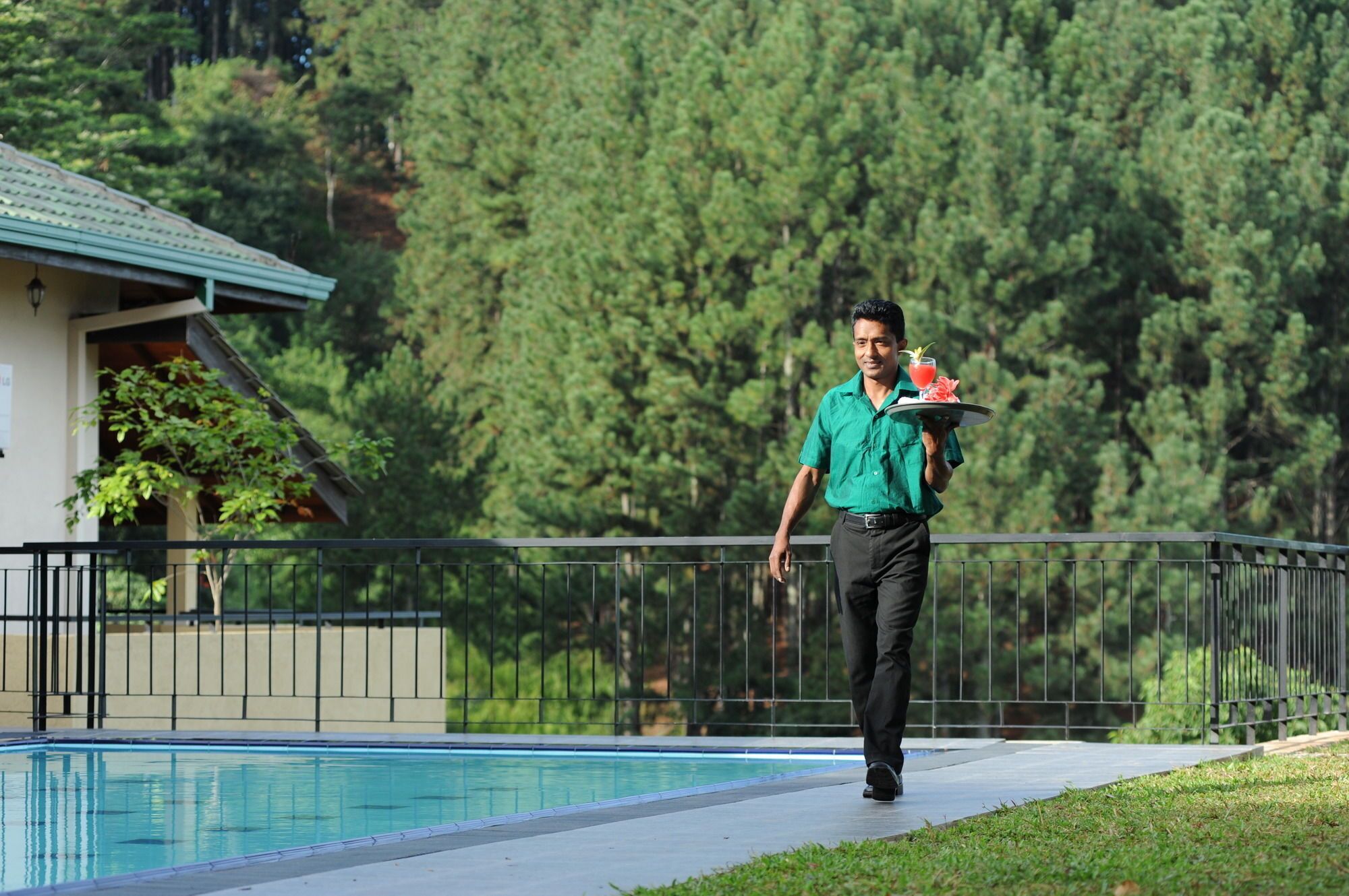 A person carrying a tray with drinks and a fruit, walking beside a pool with a house and forest in the background.