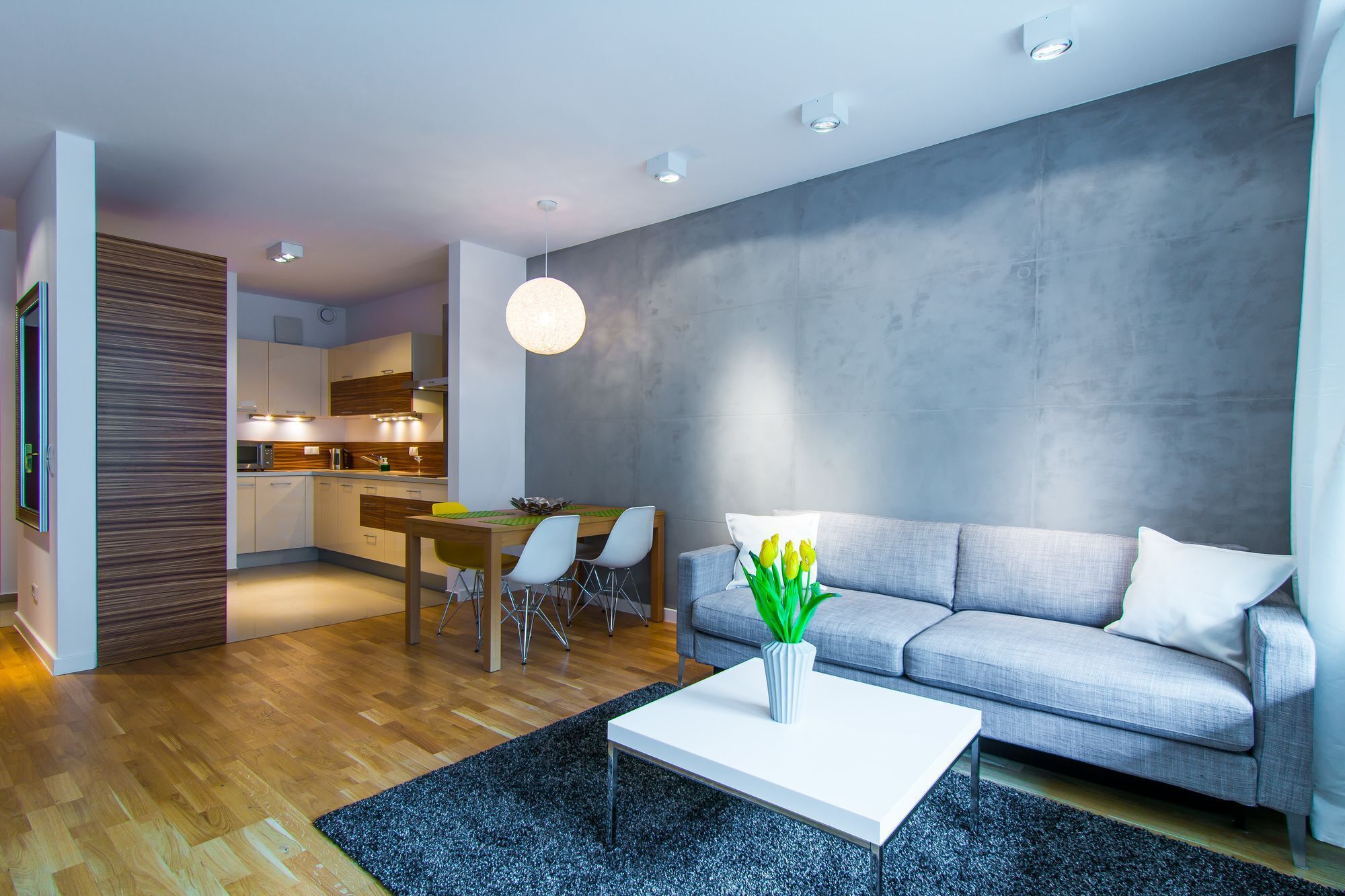 A modern living room with a grey sofa, a white coffee table, and a dining area with a wooden table and chairs.