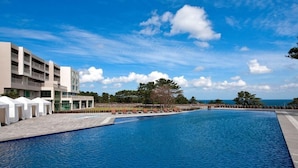 A swimming pool with lounge chairs and umbrellas, a building with multiple balconies, and a clear sky with clouds.