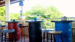 Zona de bar con taburetes y una mesa, grandes barriles azules y vistas a zonas verdes.