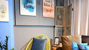 Una sala de estar con un sillón amarillo, un sofá marrón y un cojín azul. Hay cuadros enmarcados en la pared y una planta en el suelo.