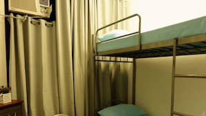 Una cama litera con un colchón individual y una almohada, un cubo de basura pequeño, un escritorio con una lámpara y una ventana con cortinas.