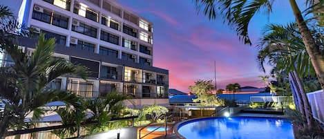 A modern hotel with a swimming pool, surrounded by palm trees and a view of the sunset.
