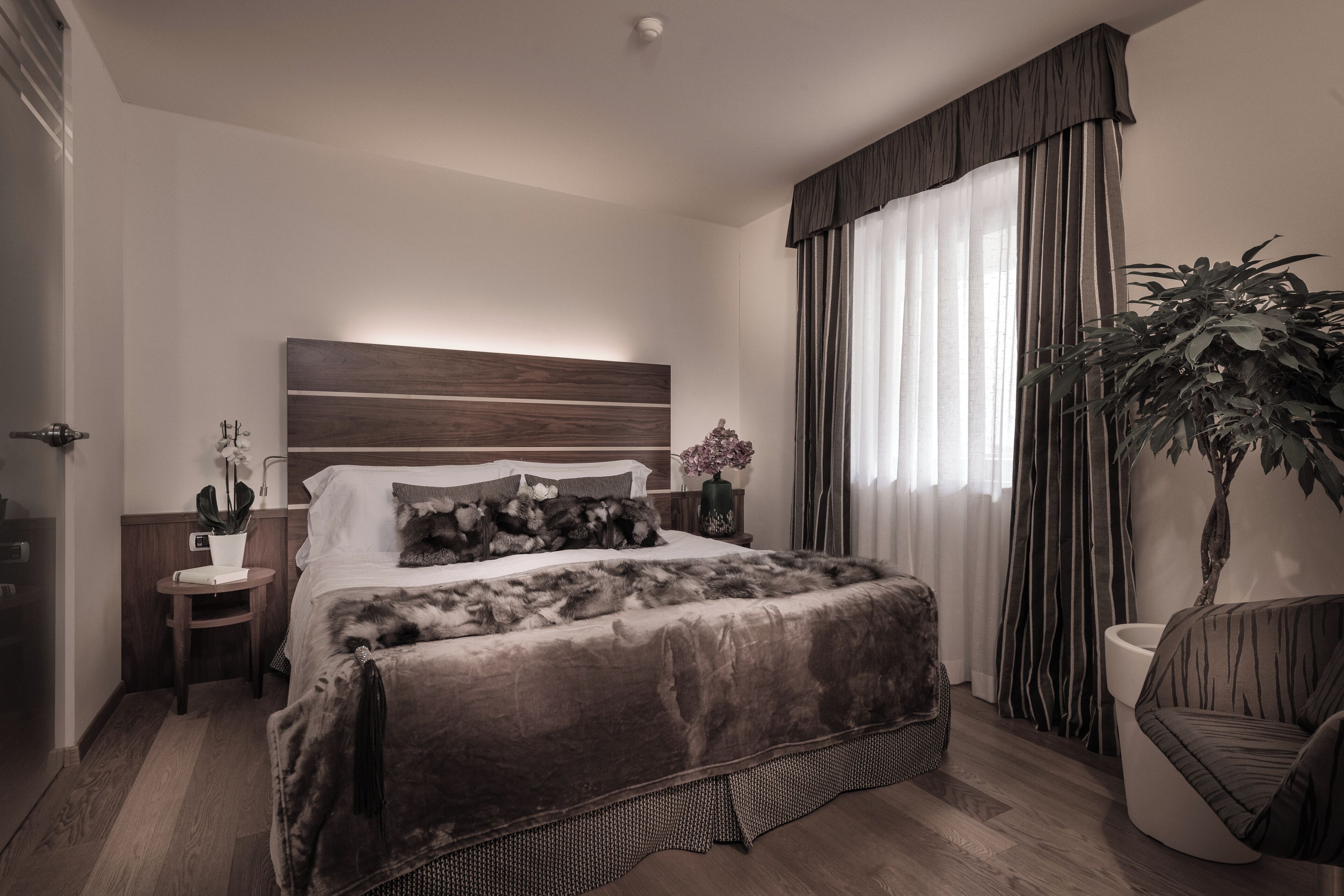 Quarto moderno com uma cama grande, mesa de cabeceira com abajur, planta em vaso e vista para o exterior através de cortinas leves.