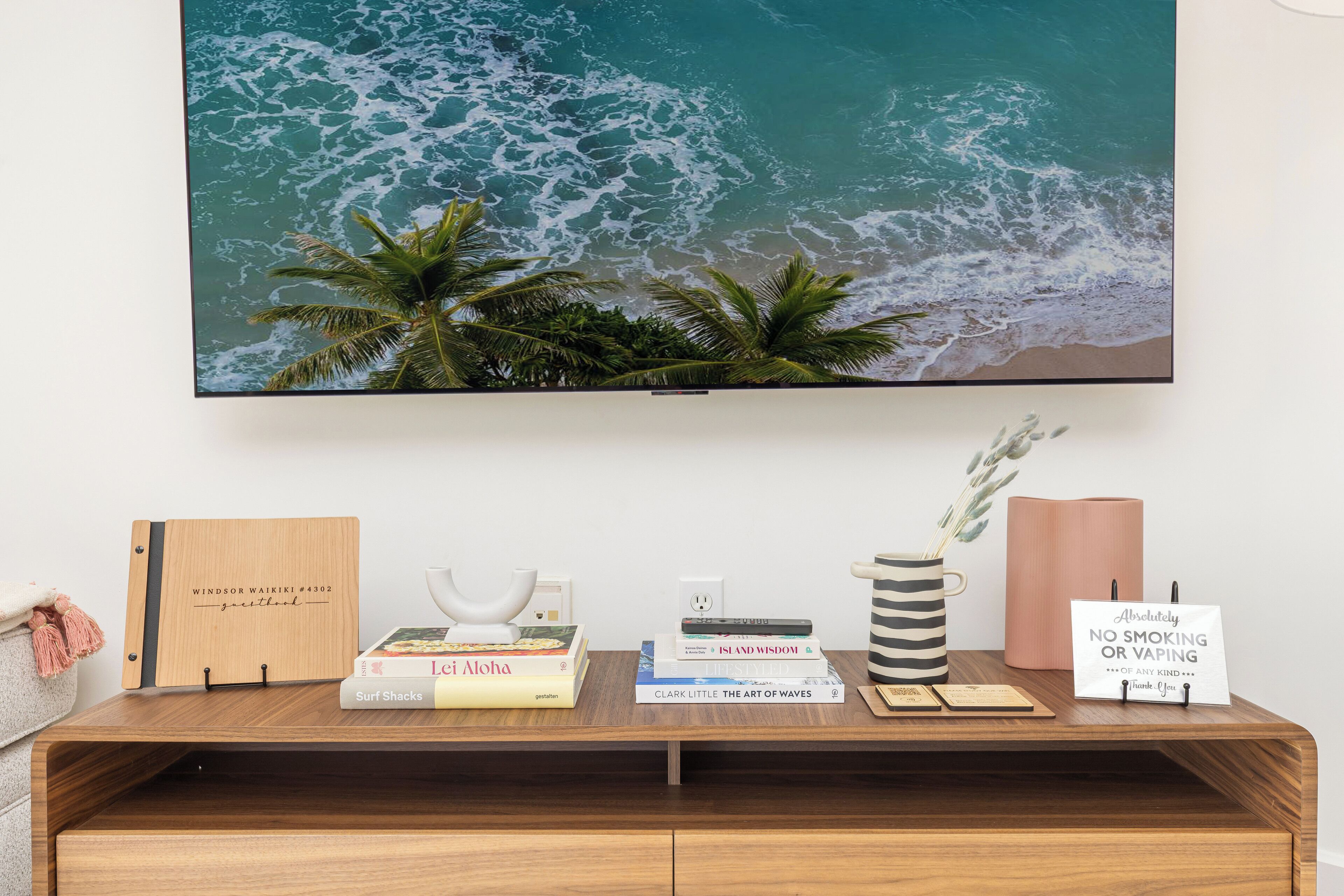 Ein Holztisch mit weitreichendem Blick auf das Meer in einem Bilderrahmen, Büchern, einer weißen Tasse, einer gestreiften Tasse, einem rosa Kerzenhalter und einem Rauchverbot-Schild.