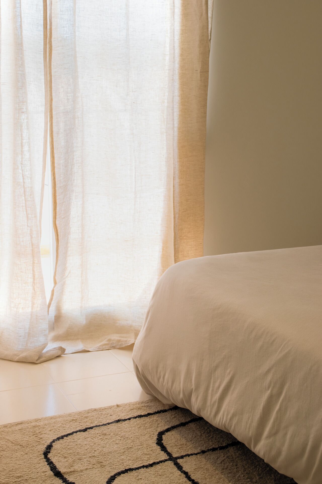 Quarto com cama, janela com cortinas leves e tapete estampado.