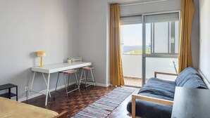 A modern living room with a white desk, two bar stools, a blue sofa, and a view of the sea through a large window with yellow curtains. - Mivô | Tejo Colours (Almada)