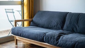 A dark blue sofa with wooden legs in a room with a wooden parquet floor and a glass door with yellow curtains. - Mivô | Tejo Colours (Almada)