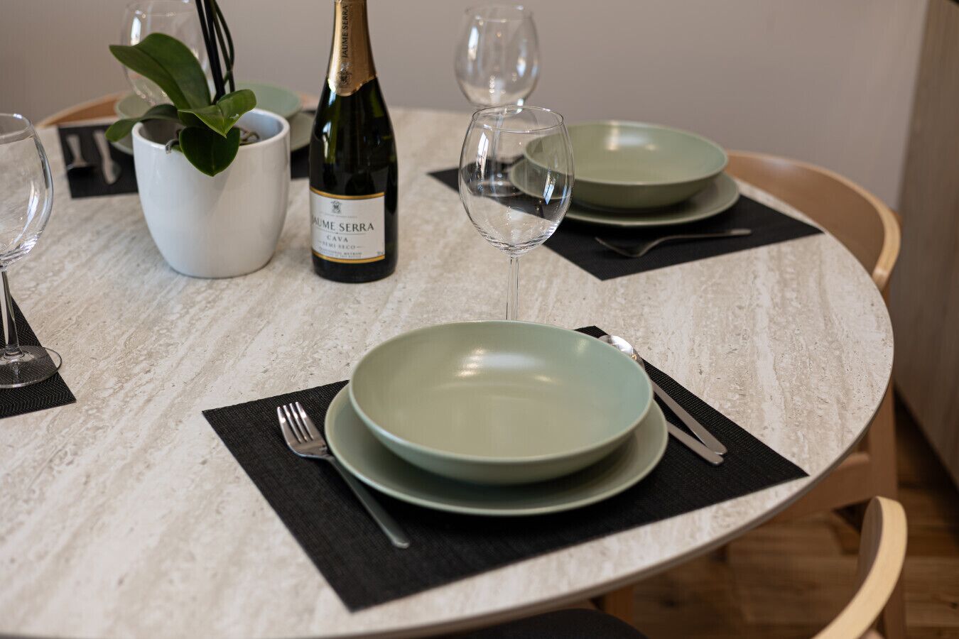 Une table à manger dressée avec de l’assiette verte, une bouteille de vin et une plante en pot.