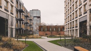 Moderni kaupunkipiha, jonka keskellä kulkee pääkävelyreitti. Pihan ympärillä on useita kerroksia sisältäviä rakennuksia, joissa on parvekkeita ja suuria ikkunoita.