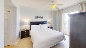 A bedroom with a bed, two bedside tables, a ceiling fan, a wall decoration, and a view of the sea. - Beachfront Charm w/ Master on Gulf 2-BR w/ Free Golf Everyday! (Panama City Beach)