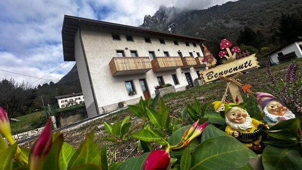 A multi-story building with balconies, a sign reading "Benvenuti," and a mountainous backdrop. - AI 7 NANI (Sedico)