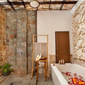 A bathroom with a stone wall, a wooden towel rack, a bathtub with rose petals, and a decorative wall carving. - Arimba Resort Ubud (Ubud)