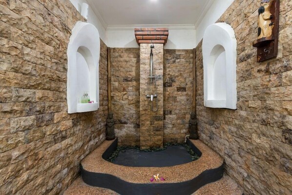 A stone-walled bathroom with a central shower area, two wall-mounted sinks, and a decorative shelf with a small plant. - Padma Retreat Ubud (Ubud)