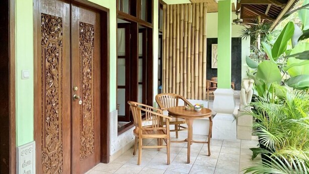 Une pièce dotée de deux portes en bois finement sculptées, d’un mur en bambou, d’un coin repas avec une table et des chaises, et de plantes luxuriantes et verdoyantes.
