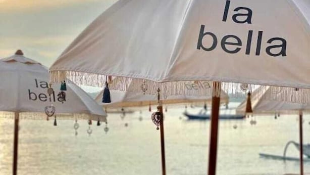 Un espace de détente aménagé sur la plage, avec plusieurs chaises longues et parasols, dont certains portent l’inscription « la bella ».