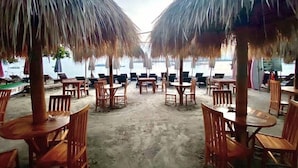 Un restaurant situé en bord de mer, avec des tables et des chaises en bois disposées sous des parasols en chaume, offrant une vue sur l’océan.