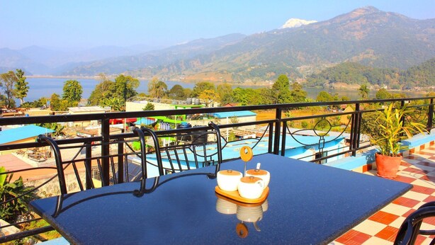 A balcony with a table, chairs, and a view of a lake and mountains. - Hotel Adam (Pokhara)