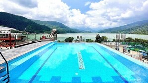 A swimming pool with clear blue water, surrounded by a deck with railings, and a view of a lake and mountains in the background. - Hotel Adam (Pokhara)