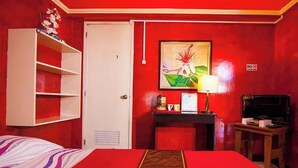 Una habitación de hotel con una pared roja, una cama con colcha a rayas, un armario blanco, un escritorio con lámpara y un televisor.