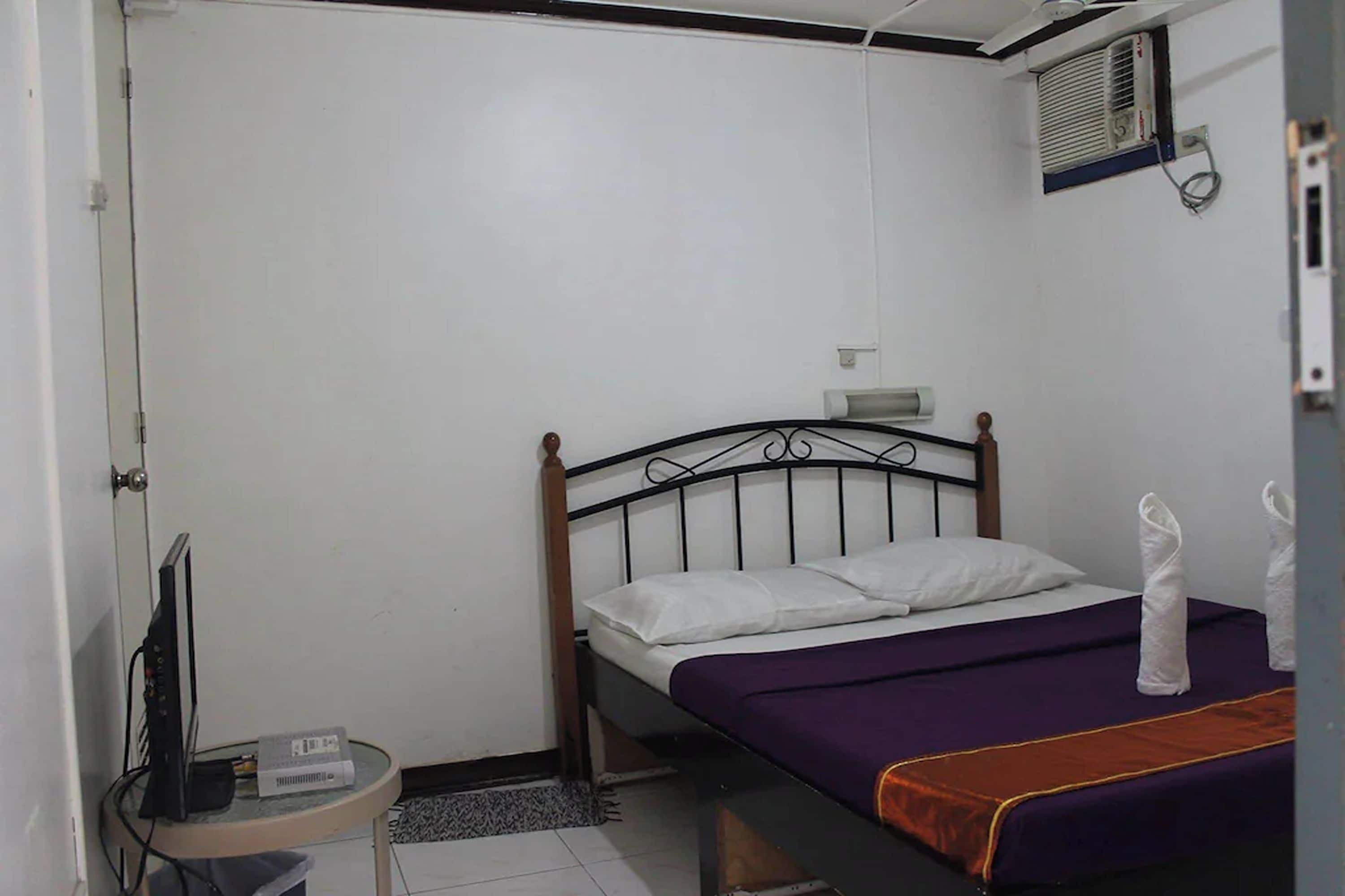A small, single-bed room with a wooden headboard, a purple bedspread, and a white pillow. There is a small table with a television and a power strip, and a wall-mounted air conditioning unit.