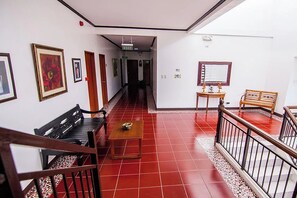 Um corredor com chão de ladrilho vermelho, bancos de madeira, uma pequena mesa e um espelho na parede.