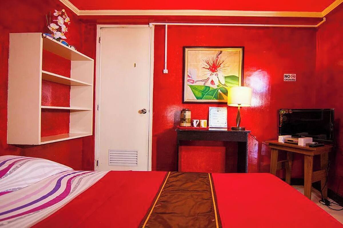 Una habitación de hotel con una pared roja, una cama con colcha a rayas, un armario blanco, un escritorio con lámpara y un televisor.