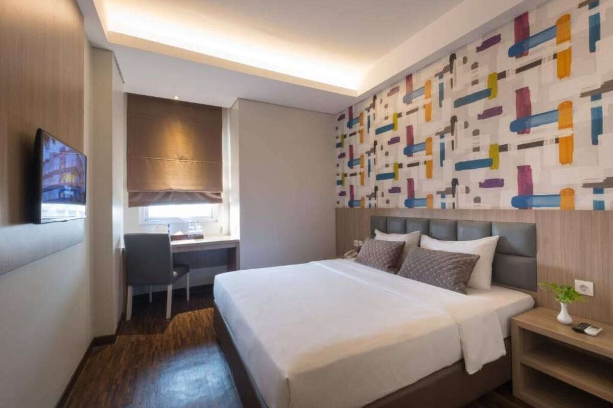 A hotel room with a bed, desk, chair, and a colorful abstract wall.