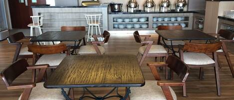 A restaurant with wooden tables and chairs, a kitchen area with appliances, and a counter with stacked plates.