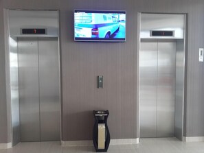 Dos ascensores con pantallas digitales, un televisor montado encima y una pequeña mesa con un florero en el vestíbulo.