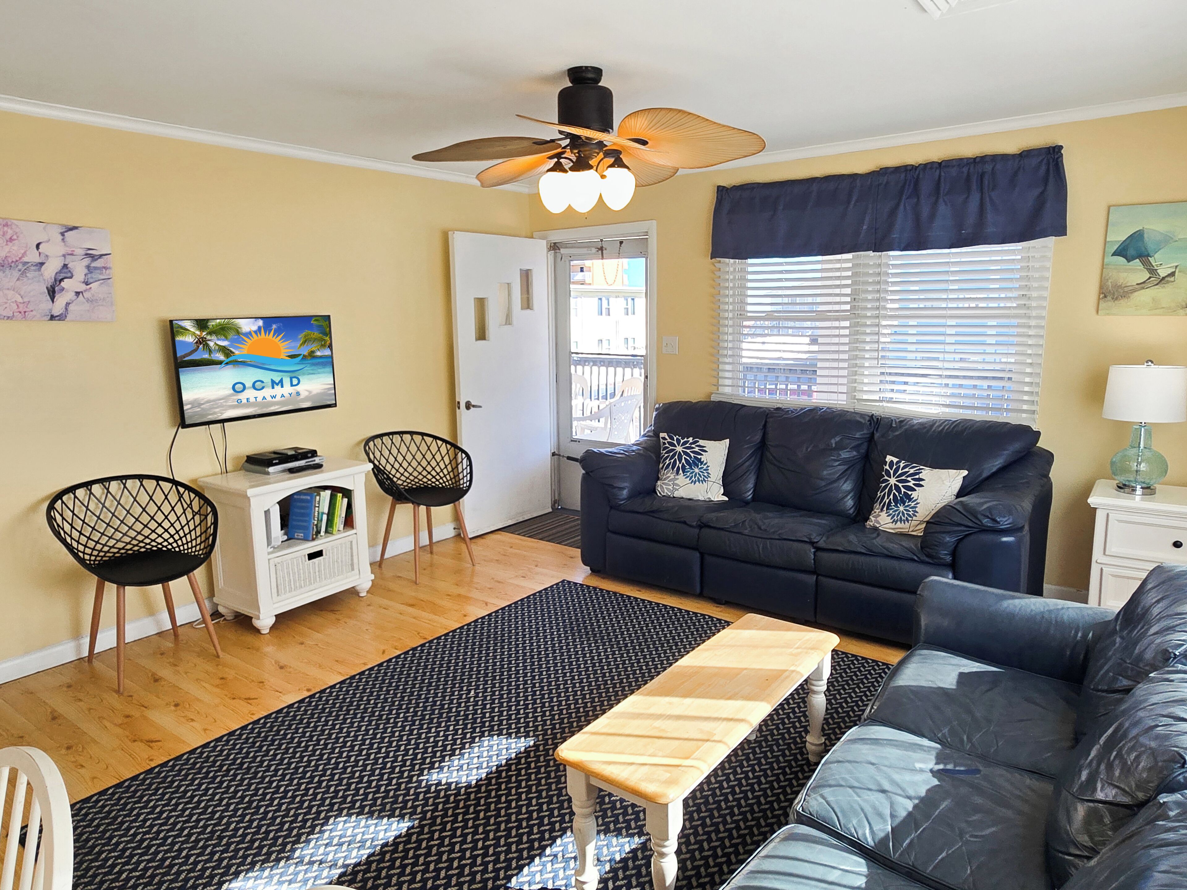 A living room with a black leather sofa, a wooden coffee table, a TV on a stand, and a ceiling fan.