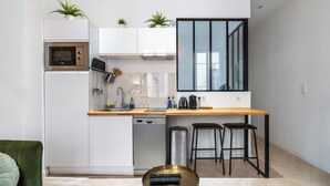 Una cucina moderna con mobili bianchi, un piano di lavoro in legno, forno a incasso, lavastoviglie e un'ampia finestra. Sono presenti due sgabelli da bar e un piccolo tavolo rotondo con un telecomando.