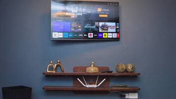 Un televisor de pantalla plana montado en la pared muestra una interfaz de servicio de transmisión con varias opciones de canales. Debajo del televisor, hay repisas de madera con objetos decorativos y una regleta de enchufes.
