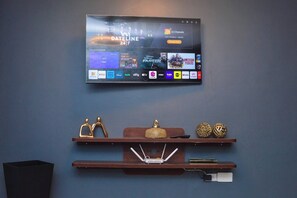Een flatscreen-tv aan een muur met een streamingdienst interface en diverse kanaalopties. Daaronder houten planken met decoratieve objecten en een stekkerdoos.