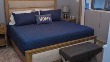 Una cama bien hecha con colcha y almohadas azules, cabecera de madera, mesita de noche con lámpara y un banco al pie de la cama.